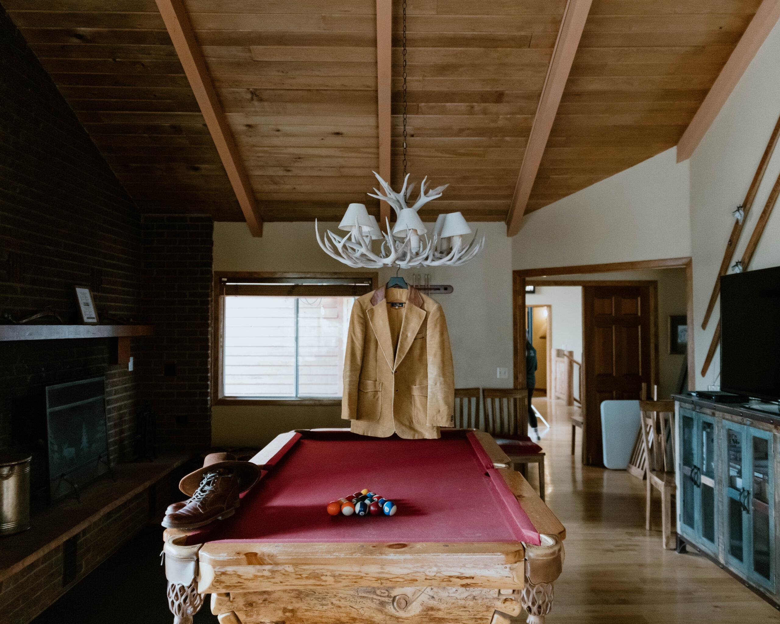 A pool table in a cabin is a resting spot for hiking boots and a hat, with an overhanging rustic chandelier