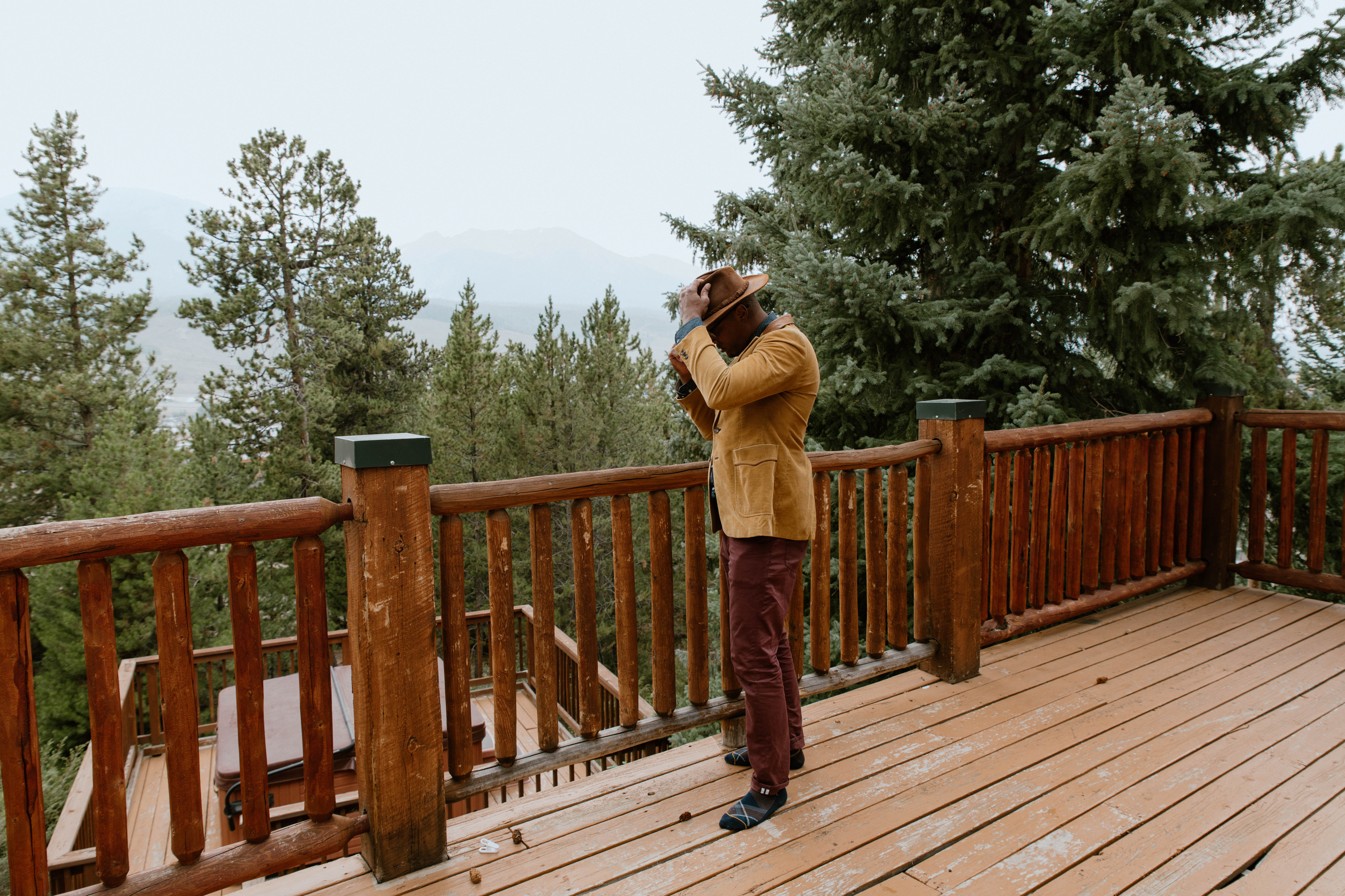 A man adjusts his hat while on a patio overlooking an evergreen forest