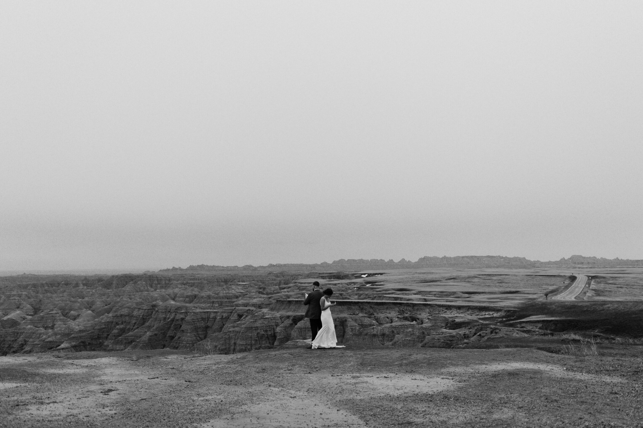 A black and white wedding photo shows a couple admiring Badlands National Park