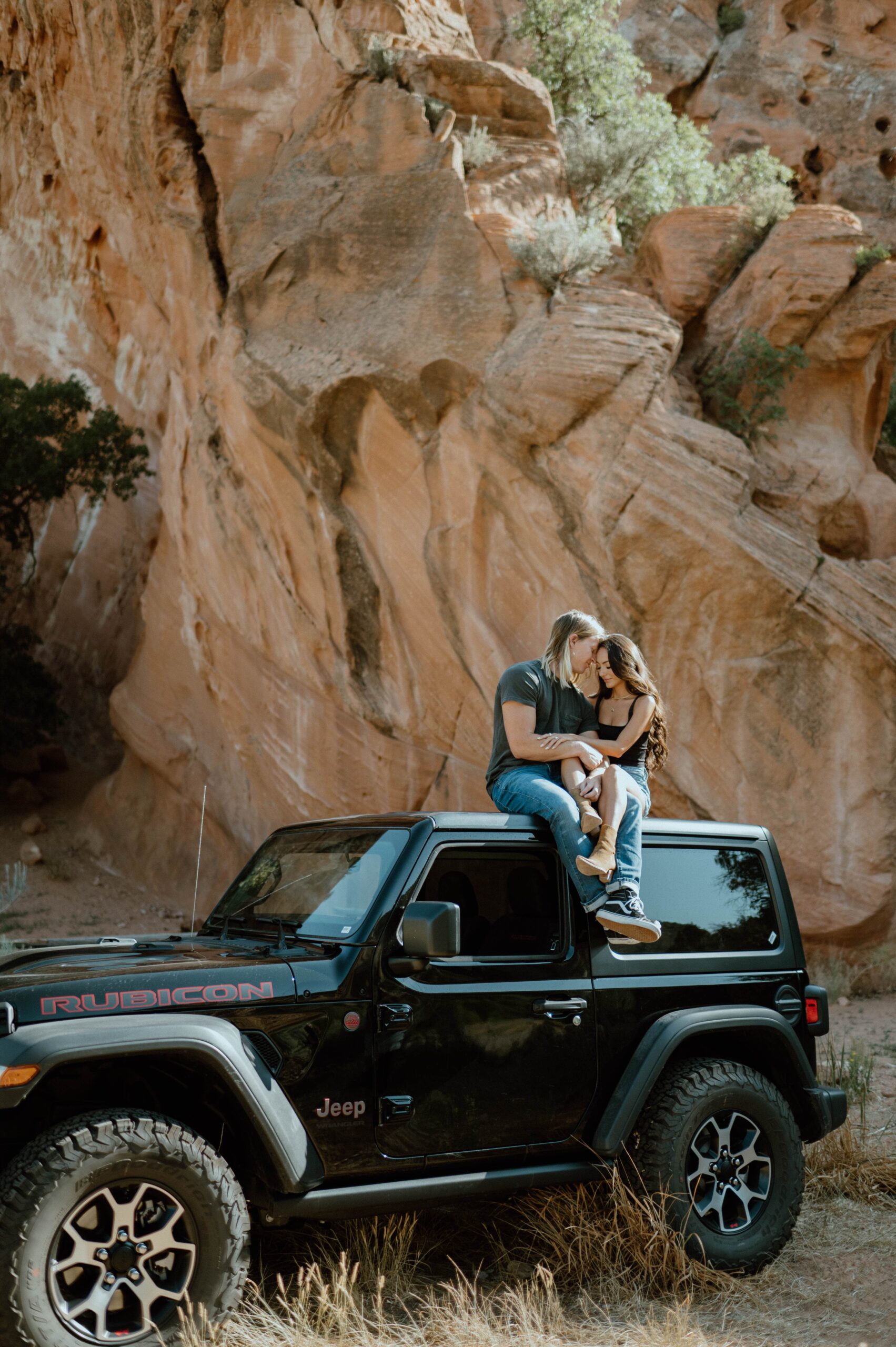 A black Jeep is pictured with a couple sitting on it's roof in Utah