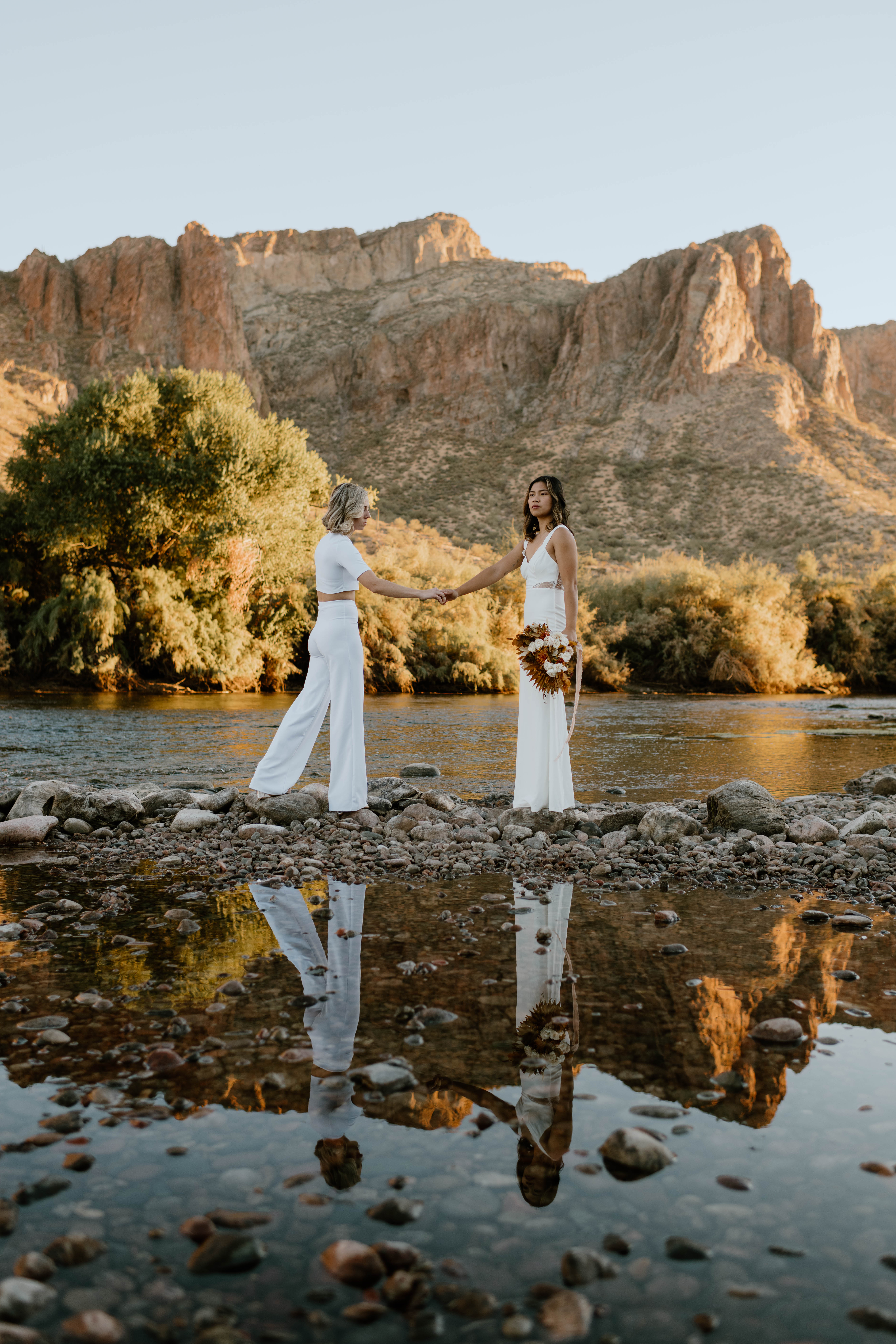 Two brides stand on a rocky water's edge in a desert in Arizona for their wedding